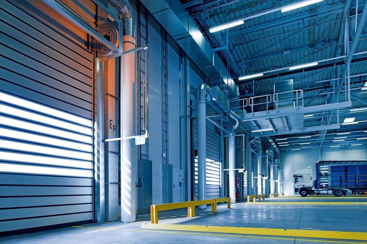 Interno di un capannone logistico con portoni industriali avvolgibili installati, spazio di lavoro illuminato con area di accesso e zona di carico e scarico.